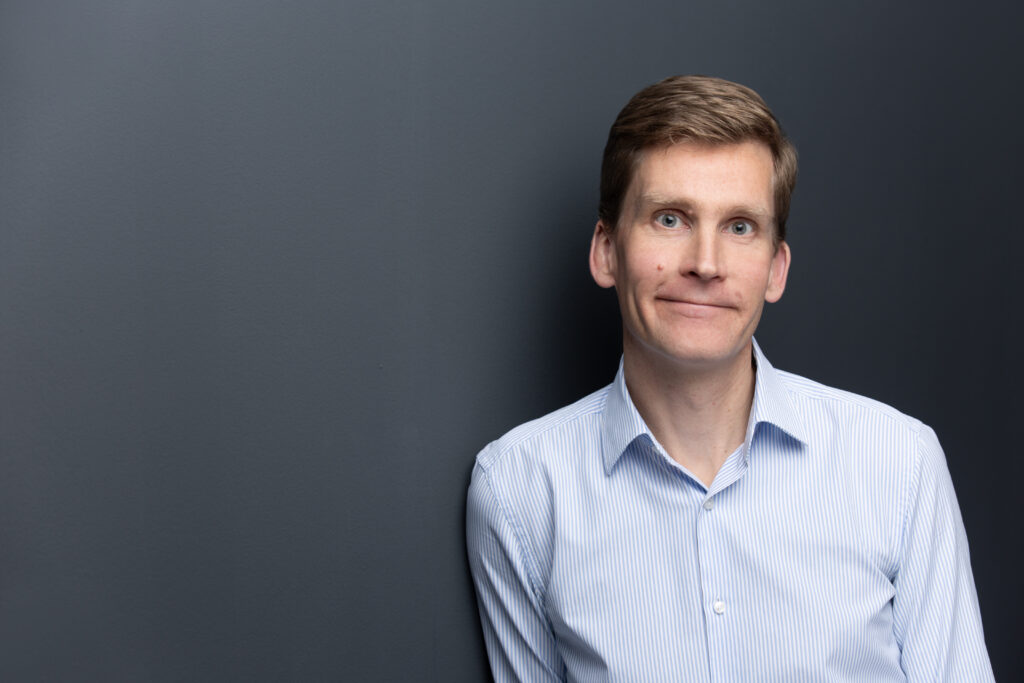 Ein blonder Mann in einem gestreiften Hemd lehnt mit der rechten Schulter an einer Wand